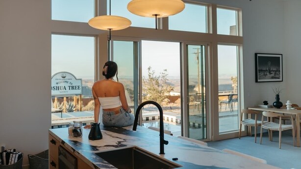 A modern kitchen with large windows, a view of the outdoors, and a sign reading "Welcome to Joshua Tree." - The Tree House By The Cohost Company (Joshua Tree)