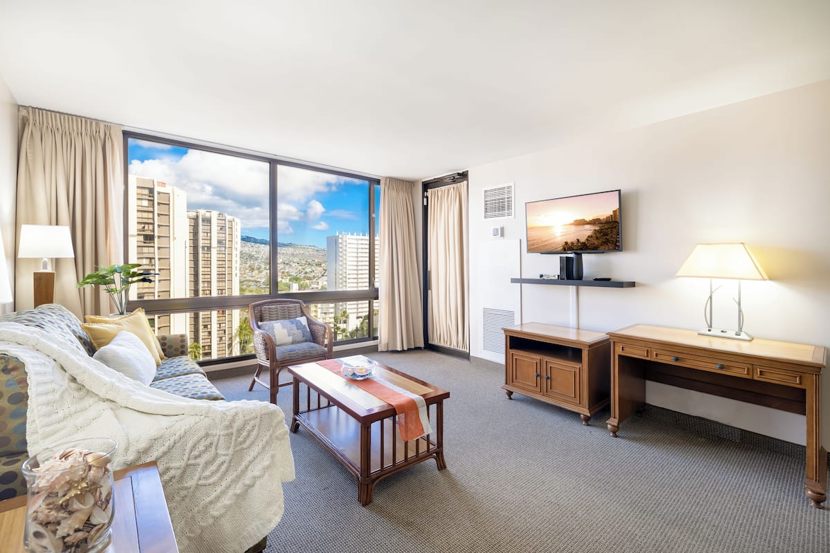 A hotel room with a large window, a sofa, a desk, a television, and a plant.