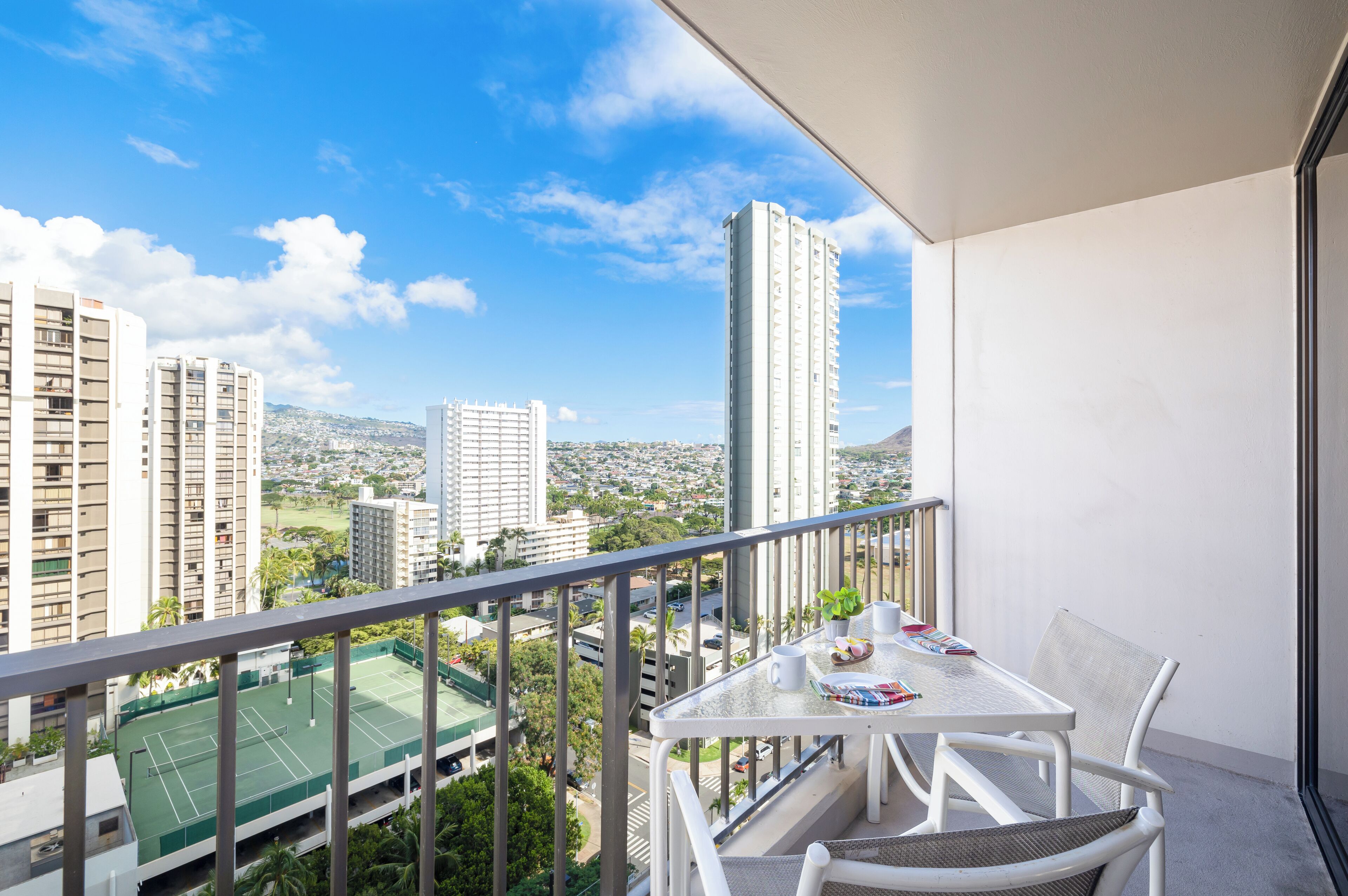 Een balkon met een tafel en stoelen, met uitzicht op een tennisbaan en stadsbeeld.