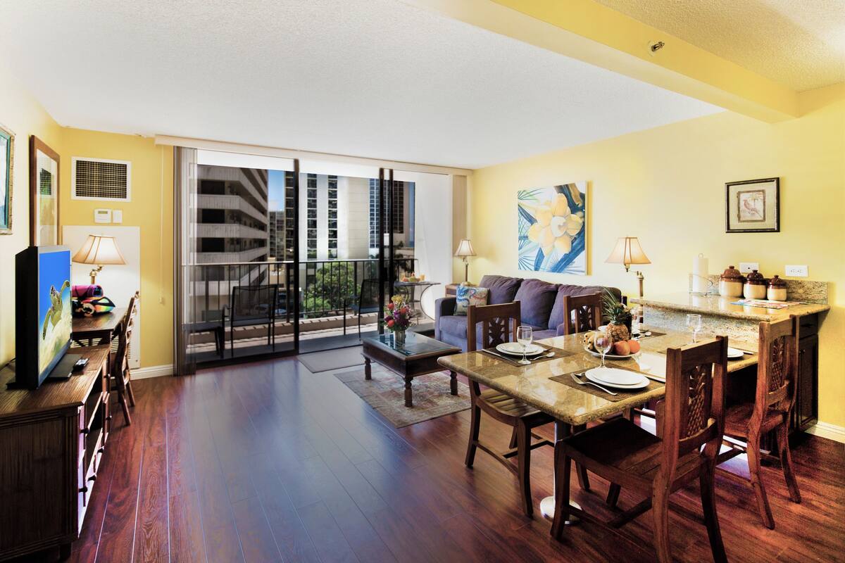 A spacious living room with a balcony, a dining area with wooden furniture, and a kitchen with a granite countertop.