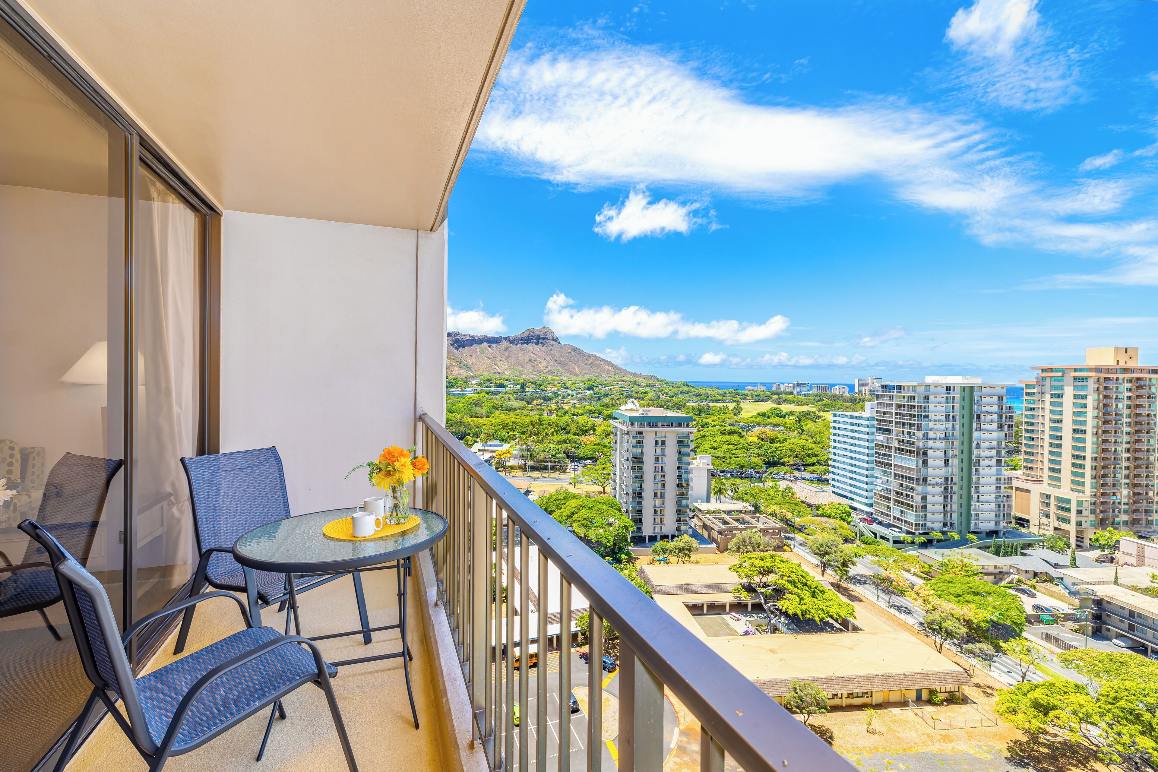 A balcony with a table and chairs, overlooking a cityscape with buildings and greenery.