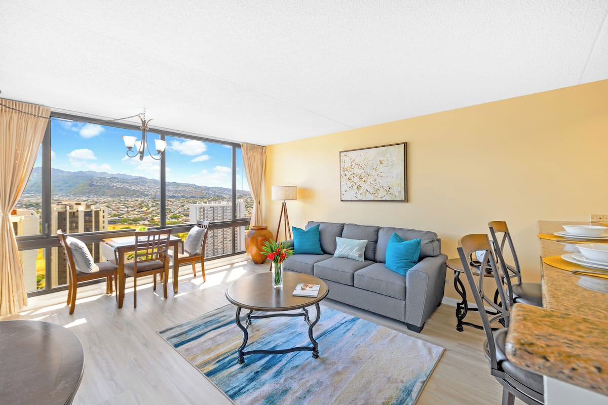 A modern living room with a large window, a grey sofa, a dining table, and a view of a cityscape.