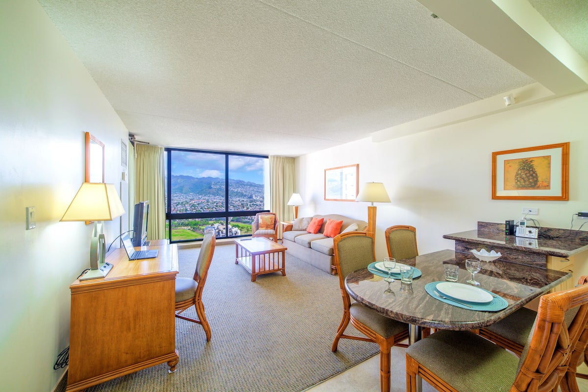 A hotel room with a dining area, a sofa, a desk, and a large window offering a city view.