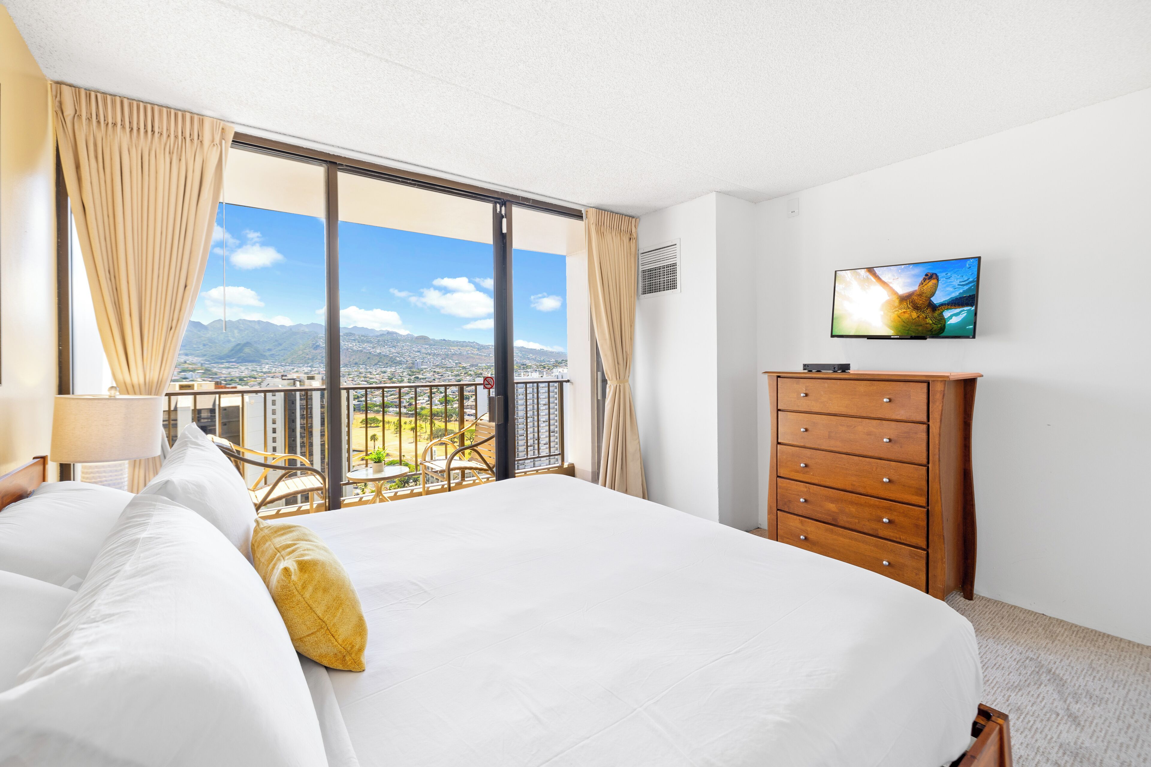 Camera d'hotel con un letto grande, un comò in legno, un televisore e un balcone con vista sulla città.
