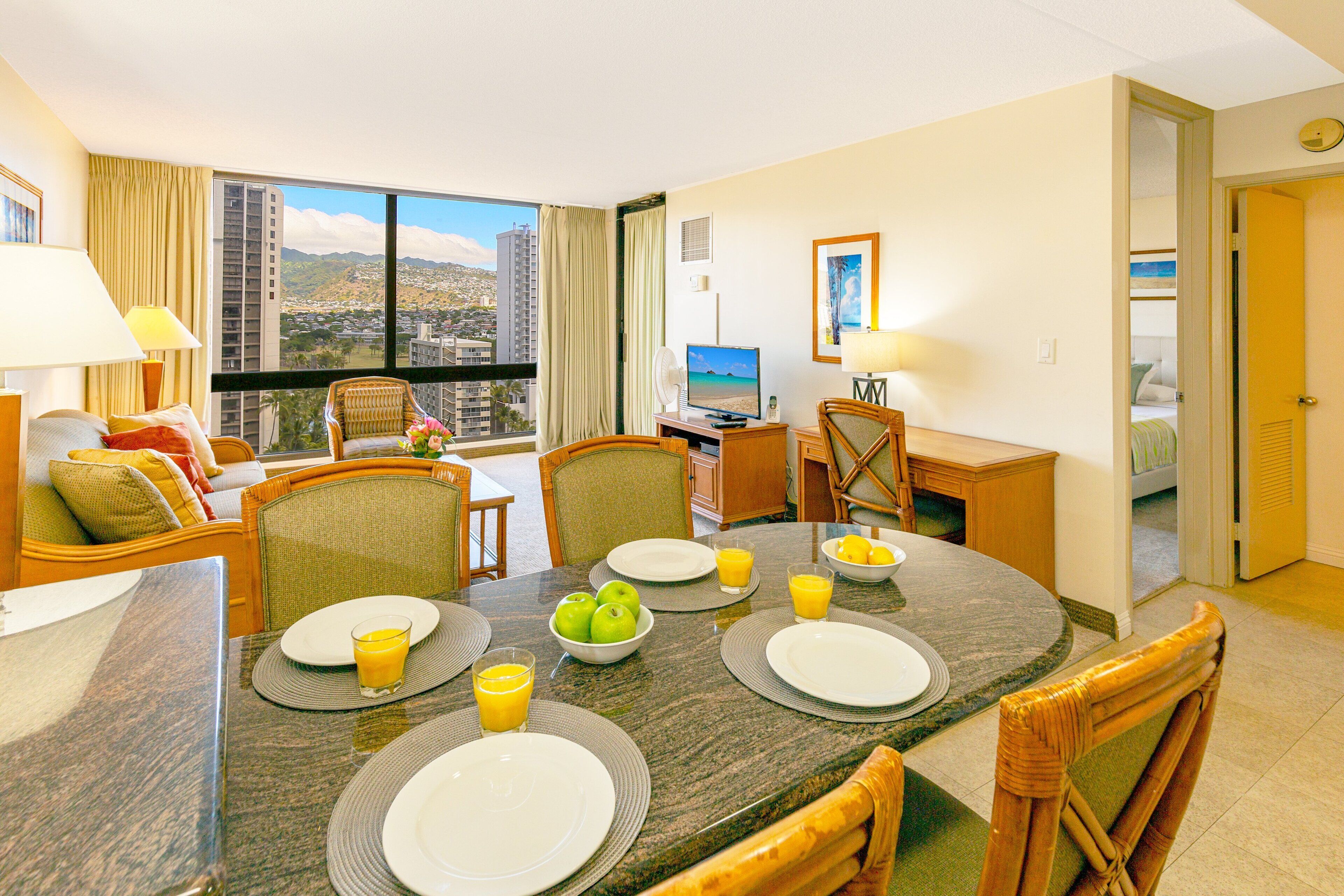 A hotel room with a dining area, a desk with a computer, and a large window offering a city view.
