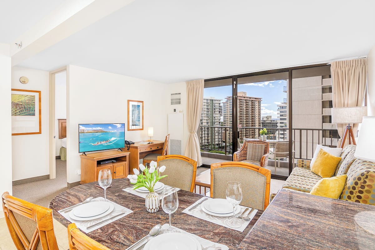A modern hotel room with a dining area, a television, and a balcony with a city view.