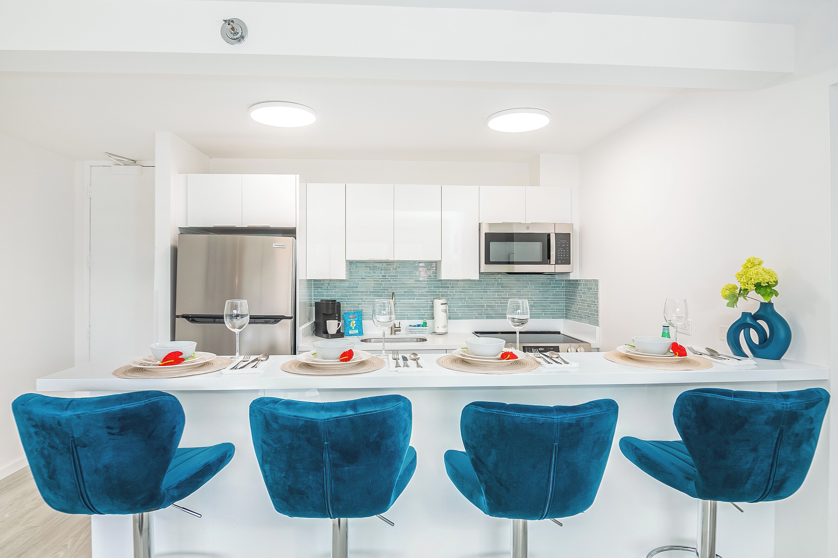 A modern kitchen with white cabinets, a stainless steel refrigerator, a microwave, and a sink. There are two blue bar stools and a white countertop with a vase of yellow flowers.