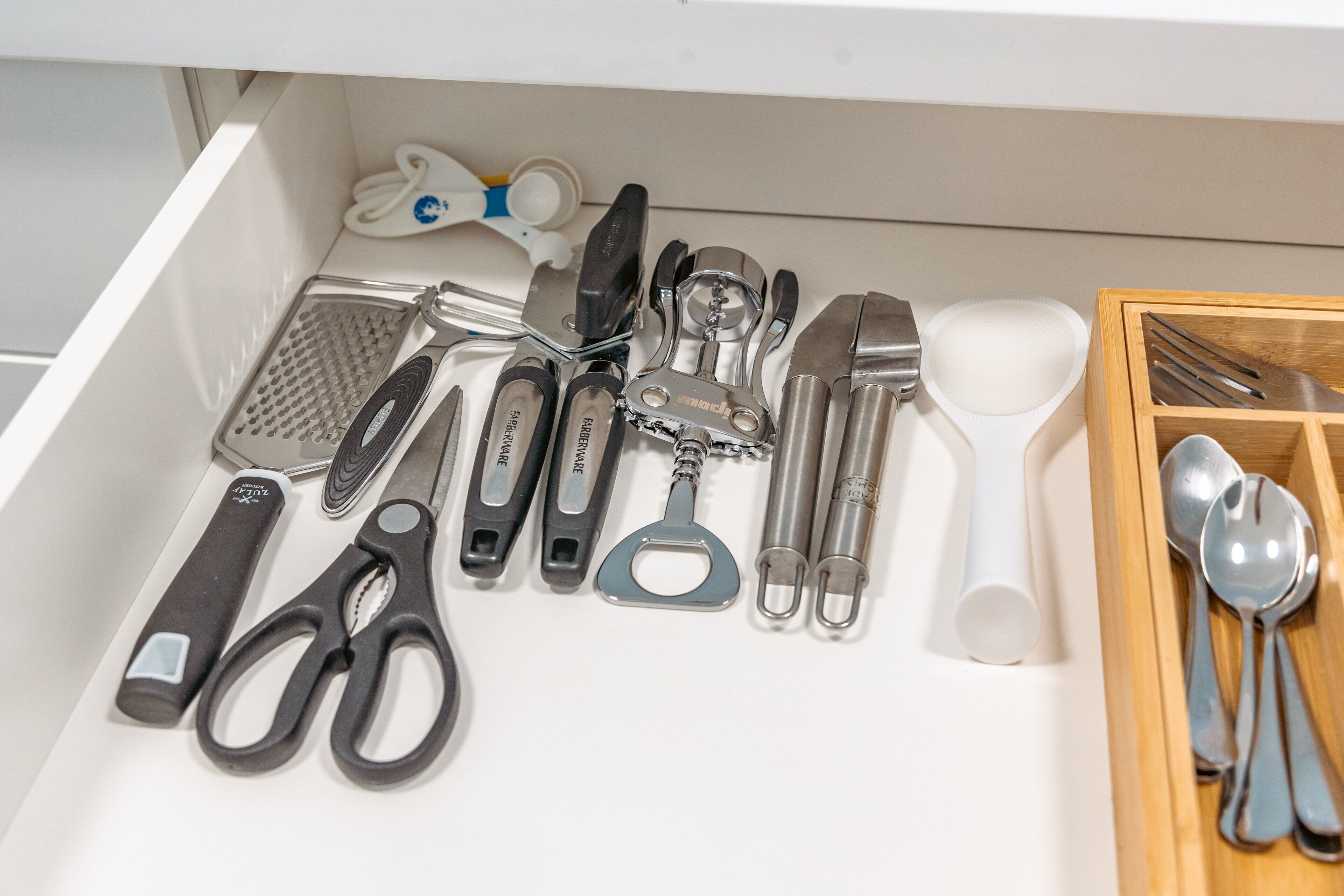 A drawer containing various kitchen utensils including scissors, a grater, a bottle opener, and a wooden spoon set.