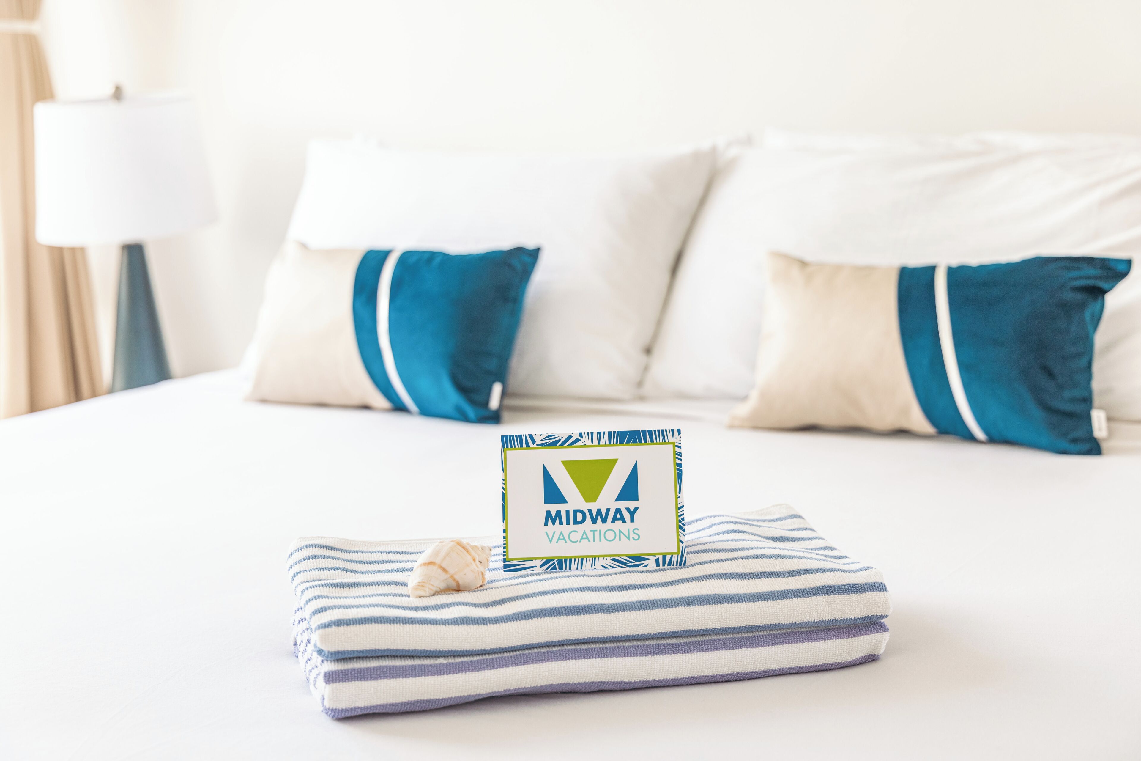 A neatly made bed with a pillow and a folded towel, a sign for Midway Vacations, and a lamp in the background.