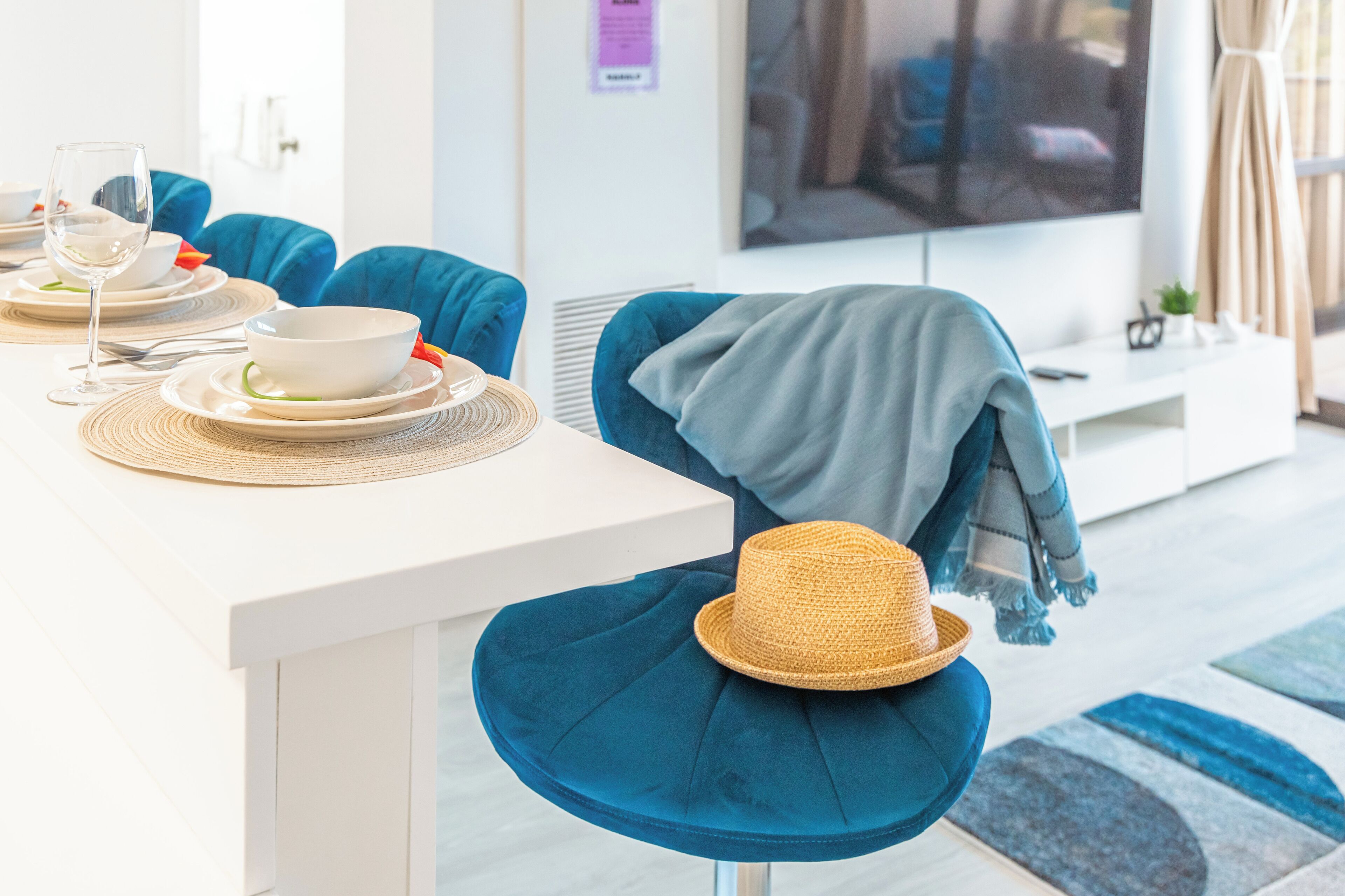 Uma sala de estar moderna com área de jantar. Há uma televisão montada na parede, uma mesa de jantar com pratos, copos e talheres. Uma cadeira azul está visível, com um chapéu de palha.
