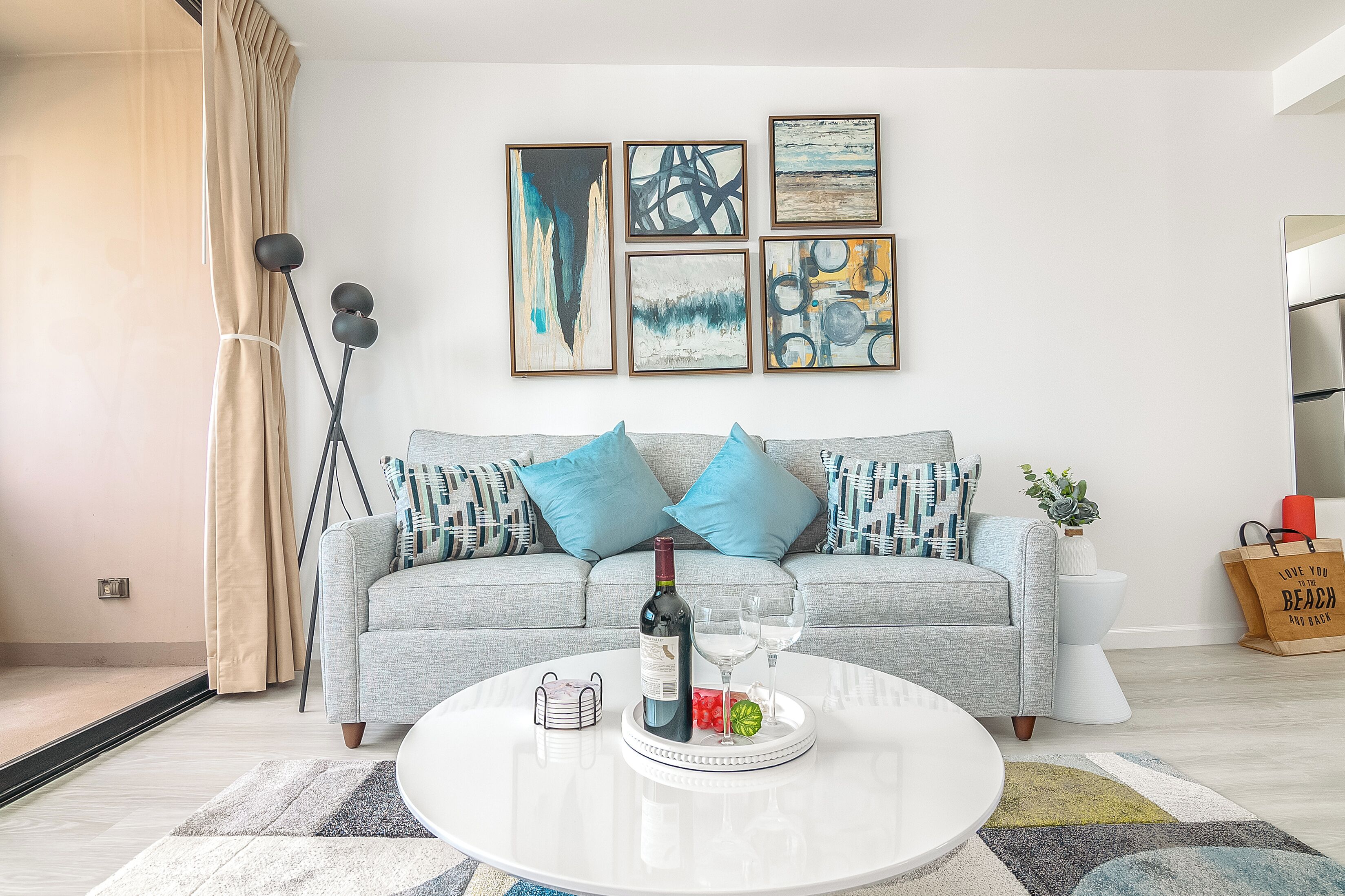 A living room with a grey sofa, blue cushions, a white coffee table with a bottle and glasses, and abstract art on the wall.