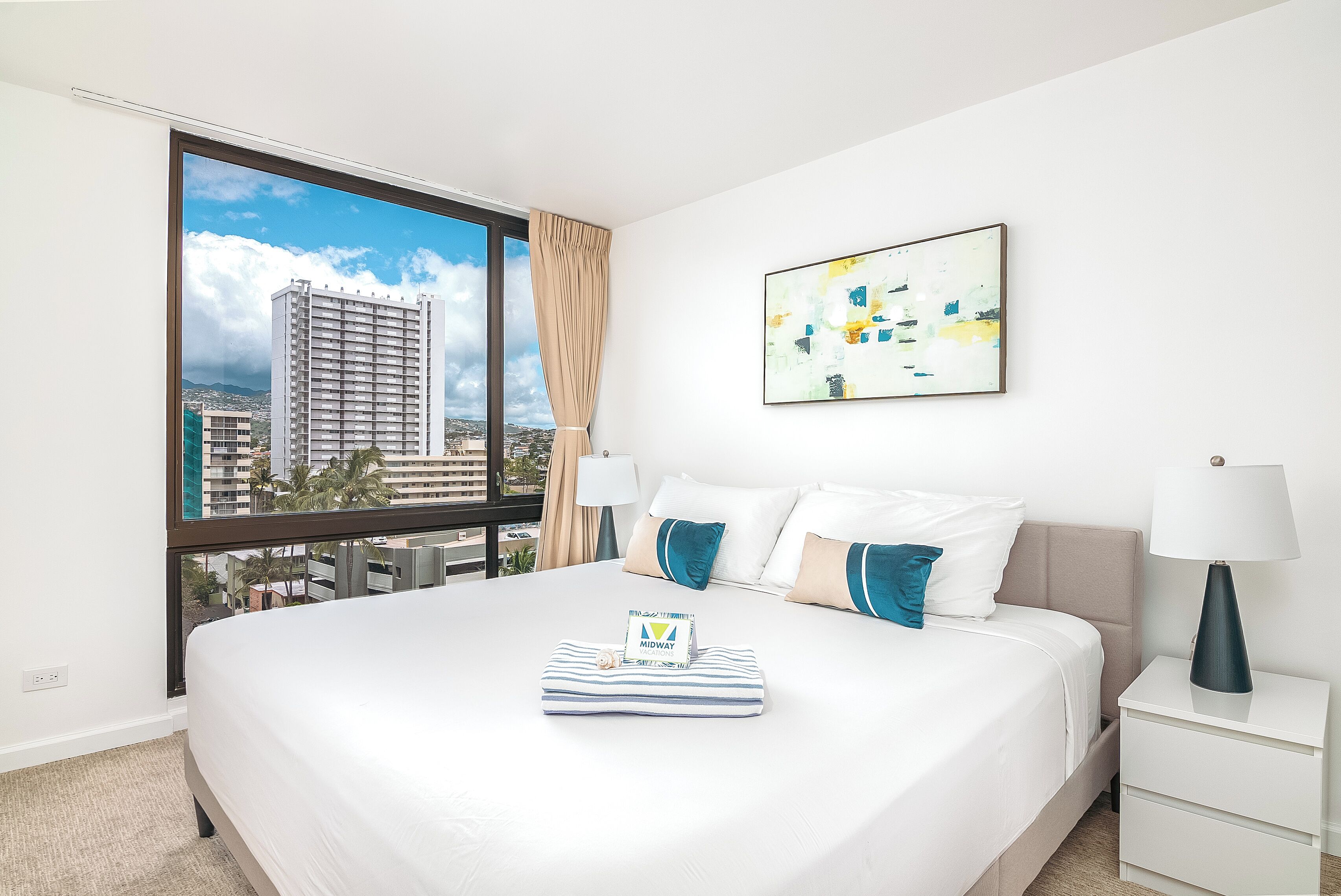 A hotel room with a large bed, a view of a cityscape, a modern abstract painting, and a neatly made bed with a pillow and a small item on it.