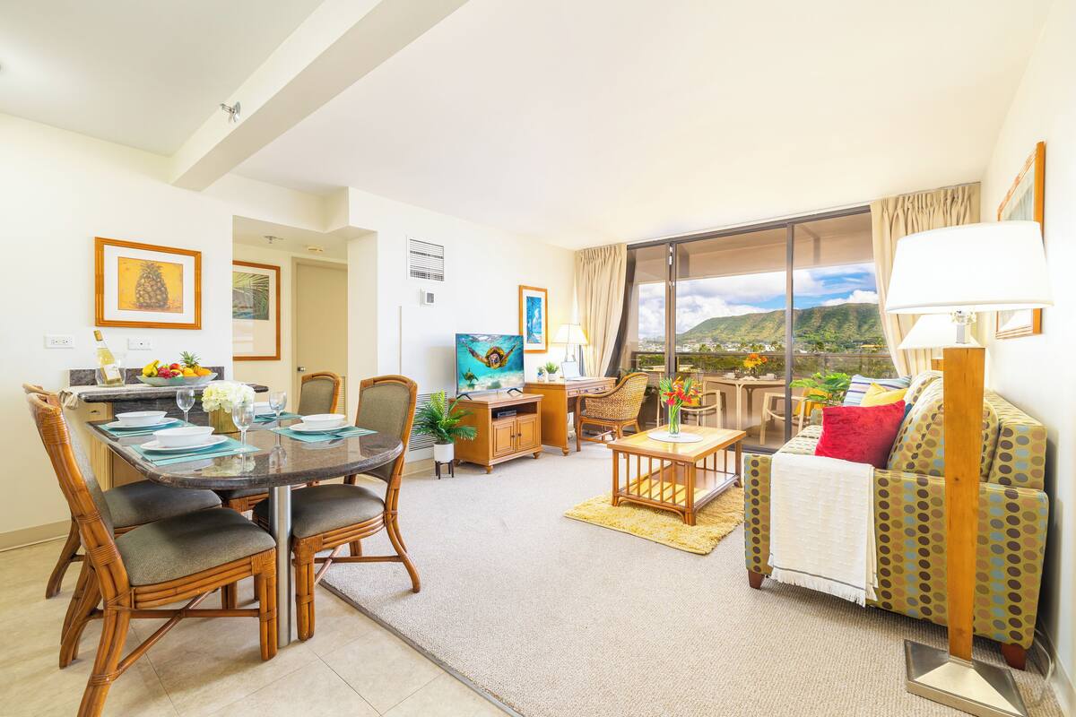 A spacious living area with a dining table, chairs, a sofa, and a view of a mountainous landscape through a large sliding glass door.