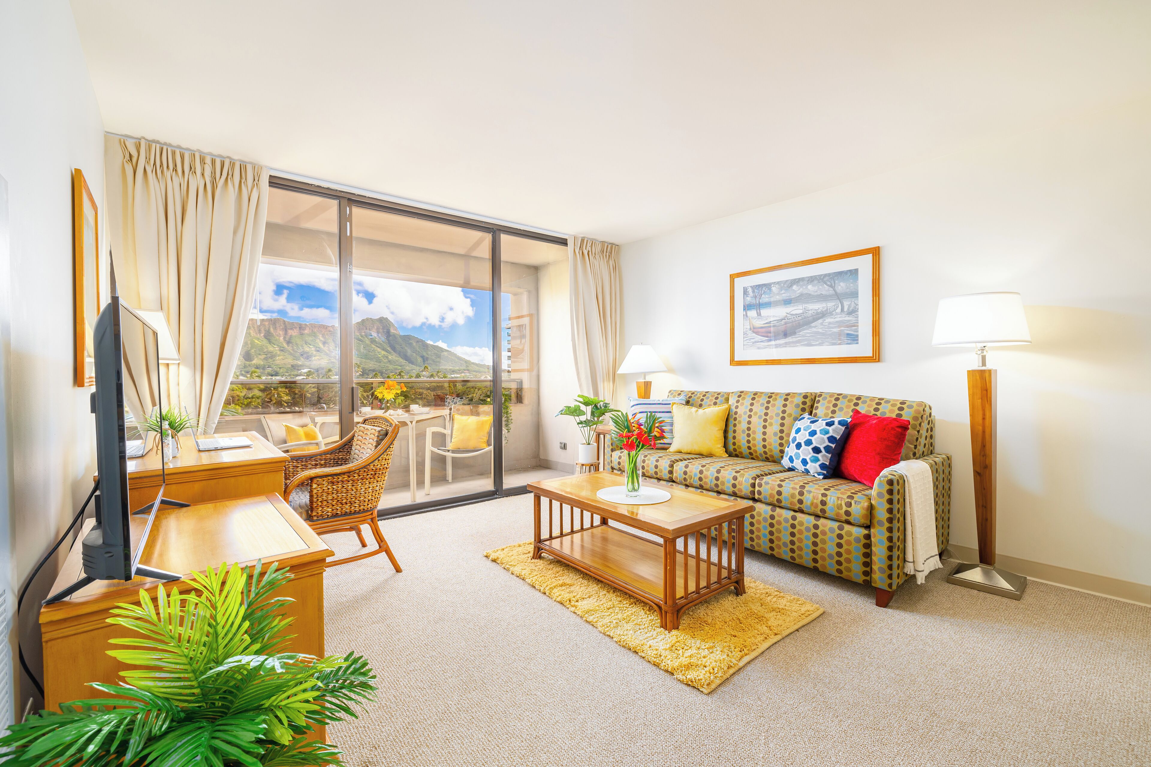 A living room with a sofa, coffee table, and a desk with a computer. There is a window with a view of mountains.