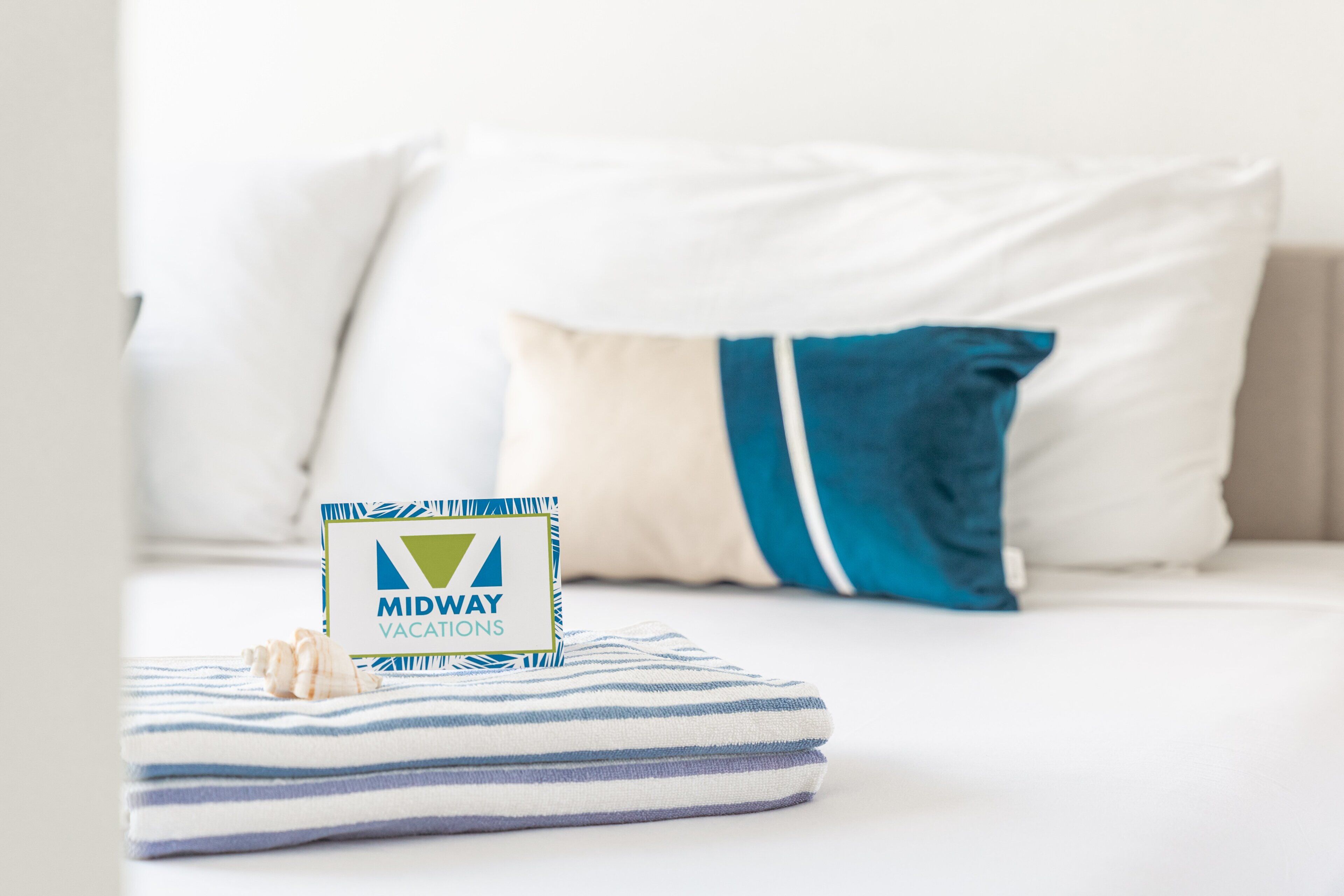 A neatly made bed with a striped blanket, a pillow with a blue stripe, and a sign for Midway Vacations.