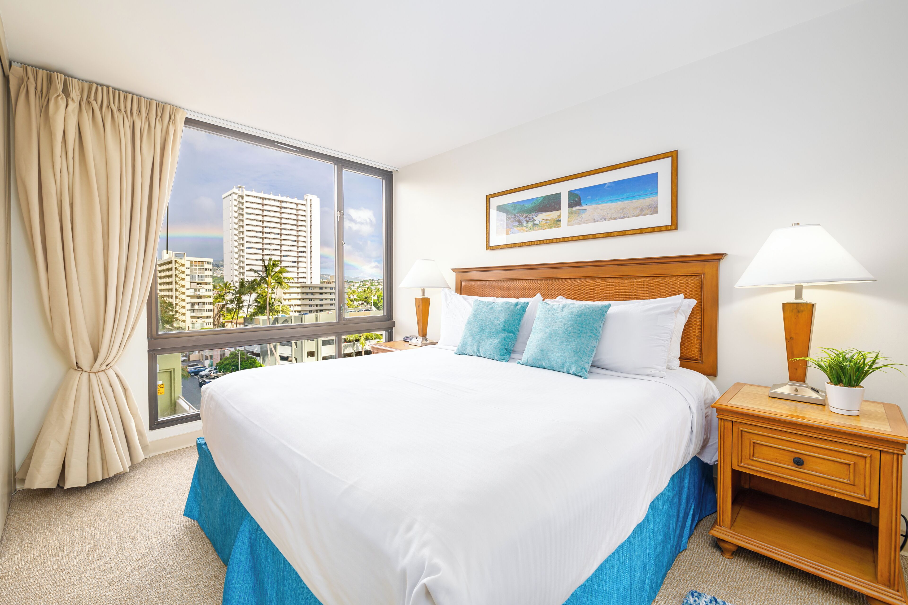 A hotel room with a large bed, a window with a view of tall buildings, a framed picture on the wall, and a nightstand with a lamp and a plant.
