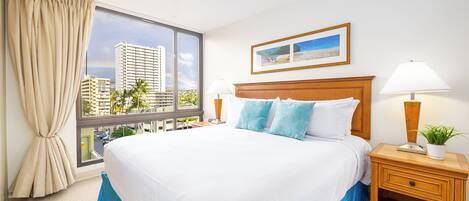 A hotel room with a large bed, a window with a view of tall buildings, a framed picture on the wall, and a nightstand with a lamp and a plant.