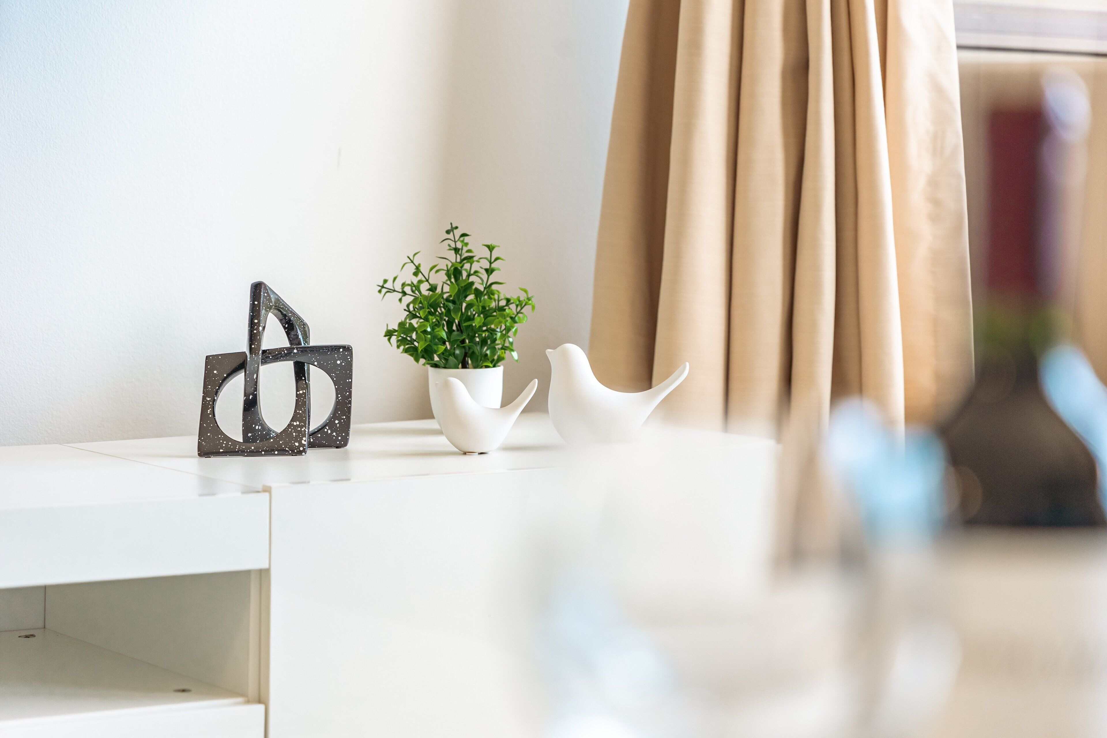 Une table blanche ornée d’une plante décorative, une paire de vases blancs en forme d’oiseaux et une sculpture abstraite noire.