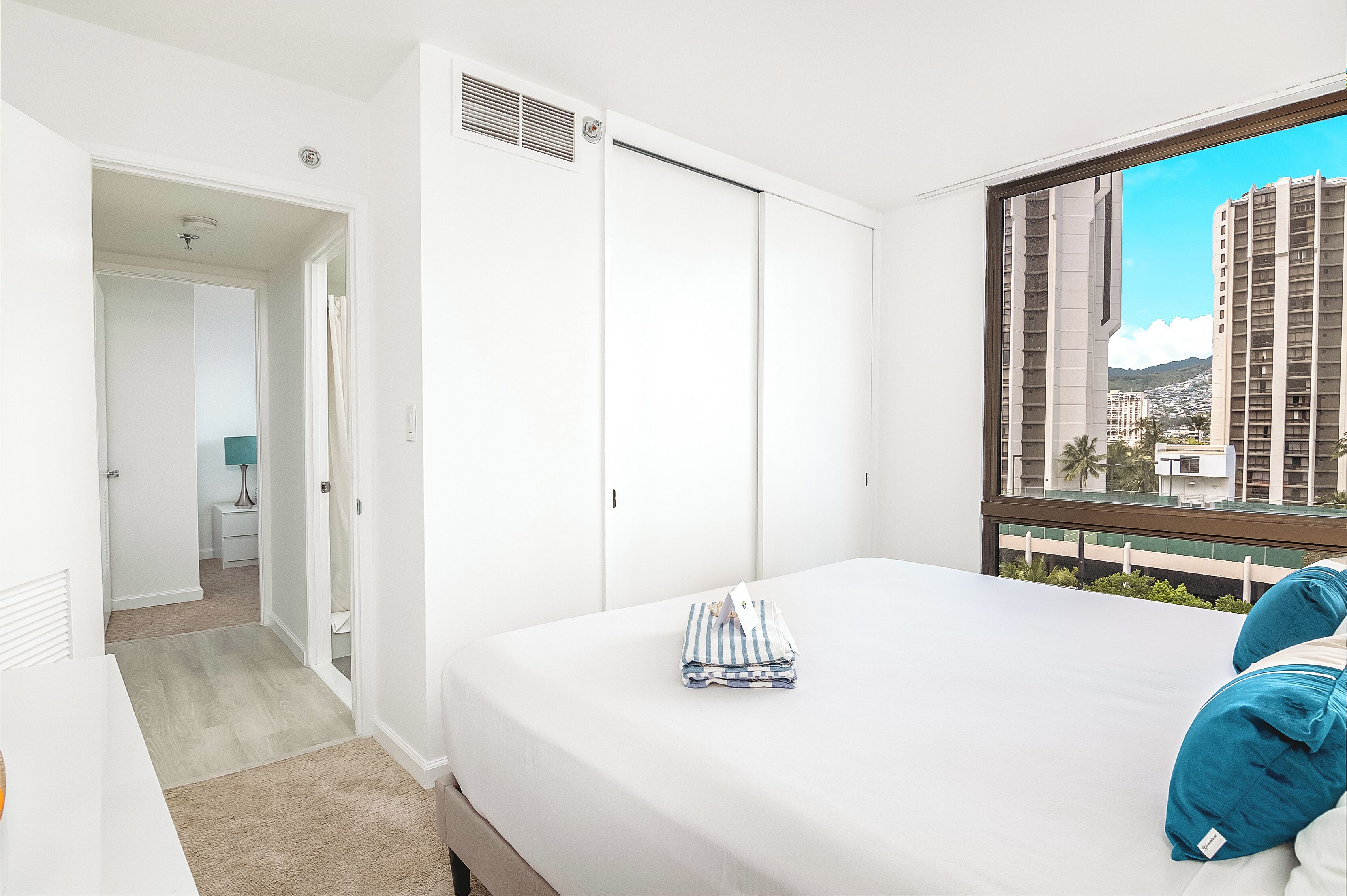 A hotel room with a large bed, a view of tall buildings, and a window with blinds.