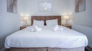 Una cama bien tendida con sábanas y toallas blancas, dos lámparas de noche y una obra de arte enmarcada sobre la cama.