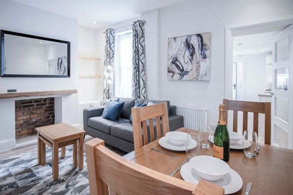 Un salon moderne avec un canapé gris, des chaises en bois, une table à manger dressée pour deux personnes et une cheminée.