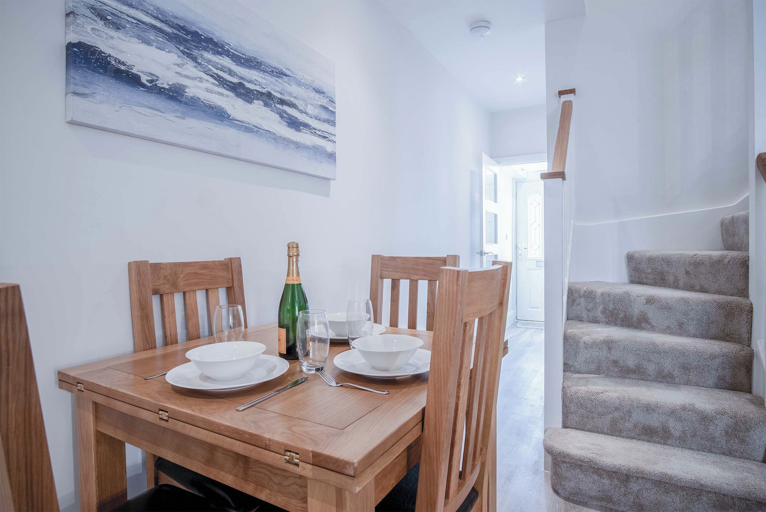 Un coin repas avec une table en bois dressée pour quatre personnes, un tableau accroché au mur et un escalier recouvert de moquette.