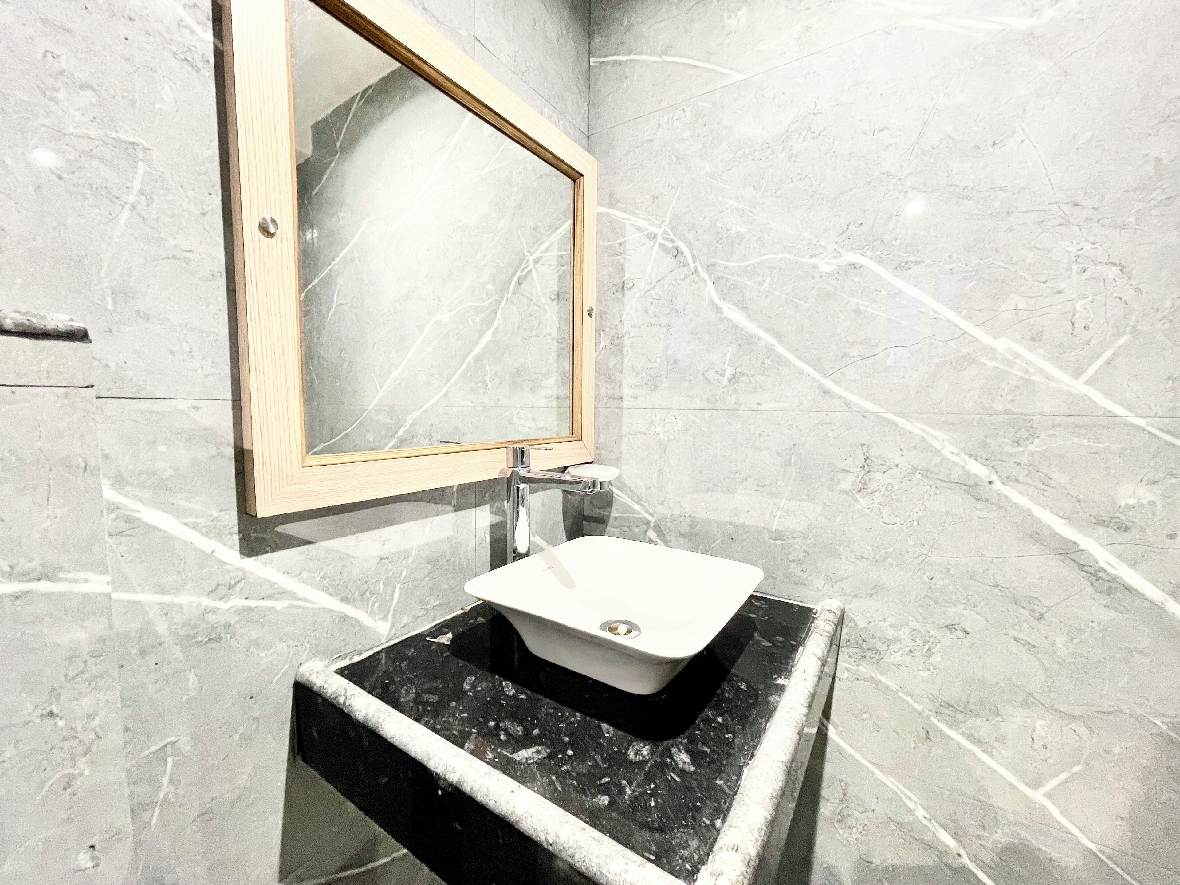 Un baño con pared de mármol, espejo con marco de madera y lavabo negro.