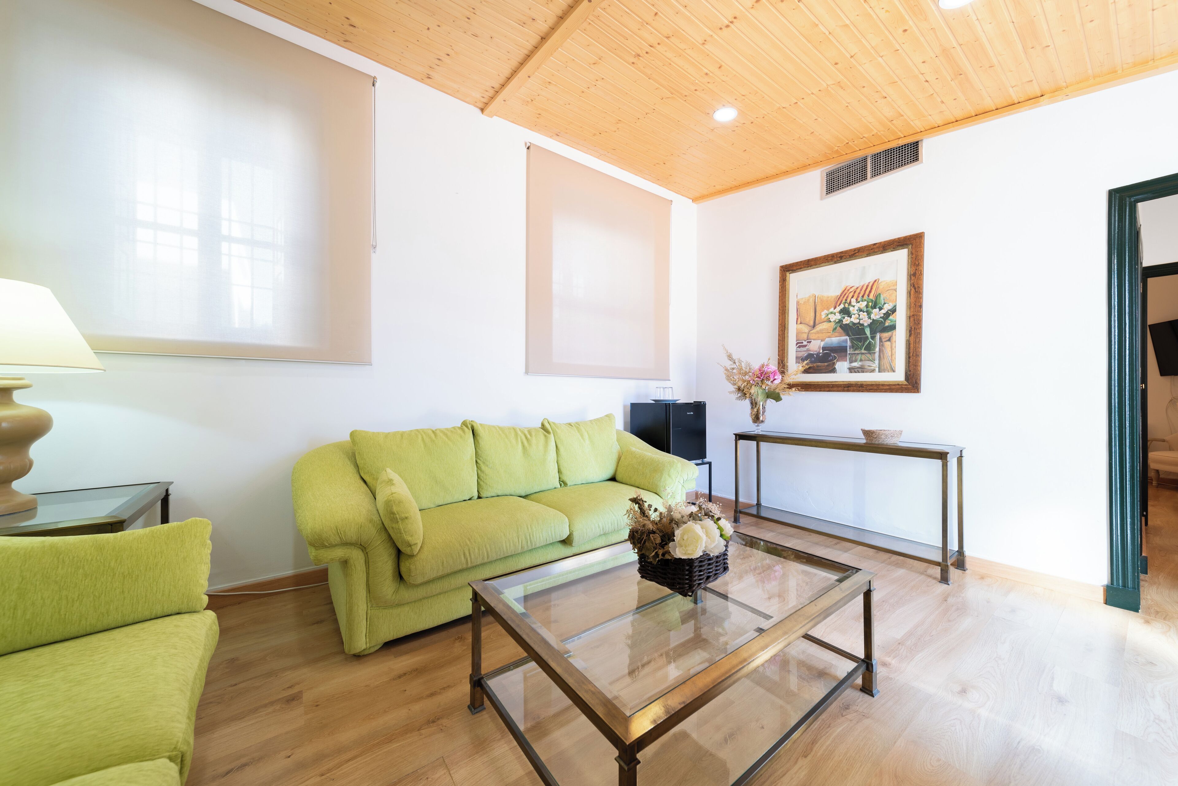 Una sala de estar con un sofá verde, una mesa de centro de vidrio, una lámpara, un cuadro enmarcado y una pequeña mesa con un jarrón.