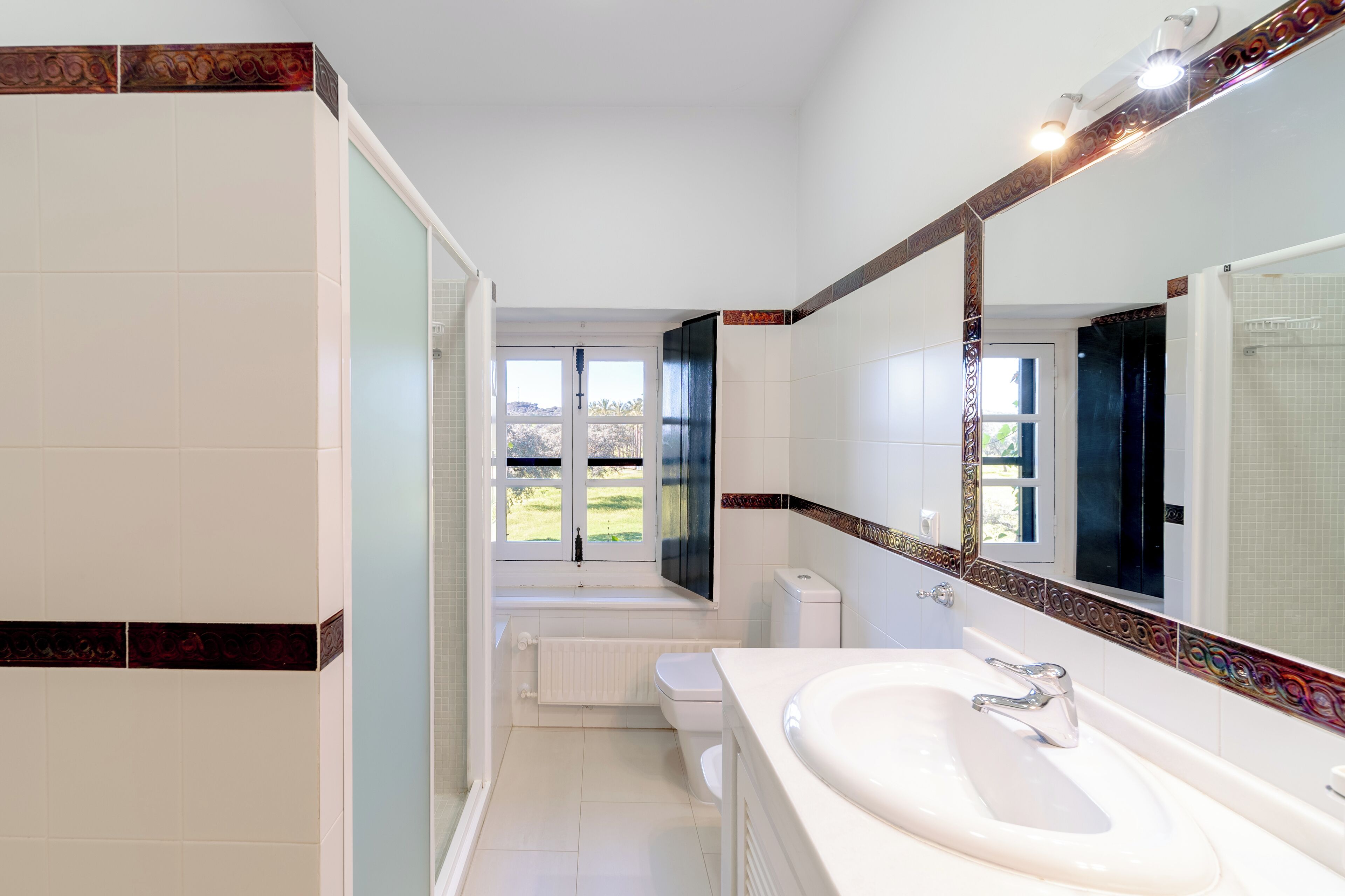 Une salle de bain moderne avec un grand miroir, un lavabo blanc et une vue sur l’extérieur par une fenêtre.