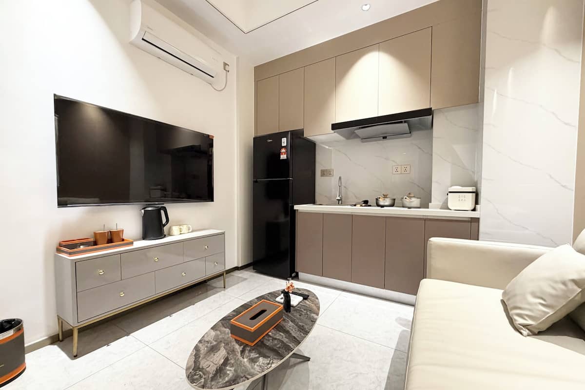 Cozinha moderna com forno embutido, micro-ondas e pia. Há uma TV de tela plana na parede, uma pequena mesa de jantar com cadeiras e um sofá na sala de estar.