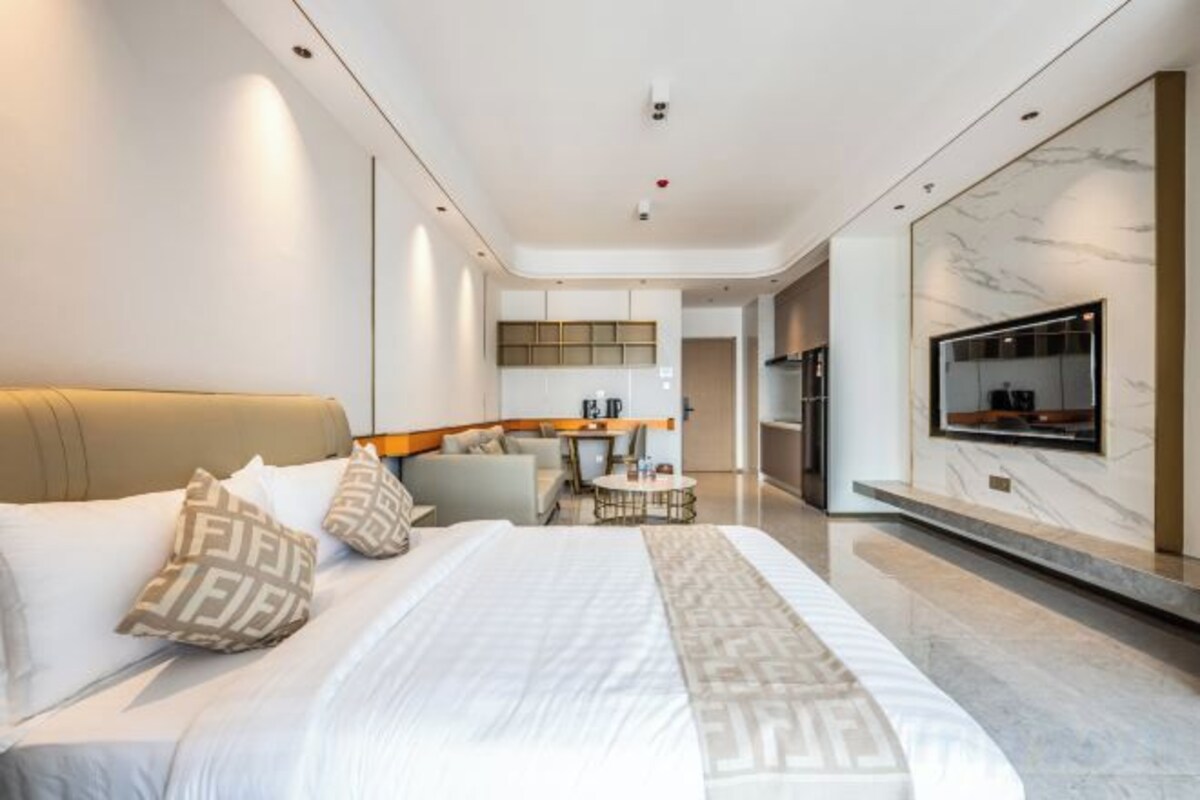 A modern hotel room with a large bed, a seating area, a TV, and a kitchen in the background.