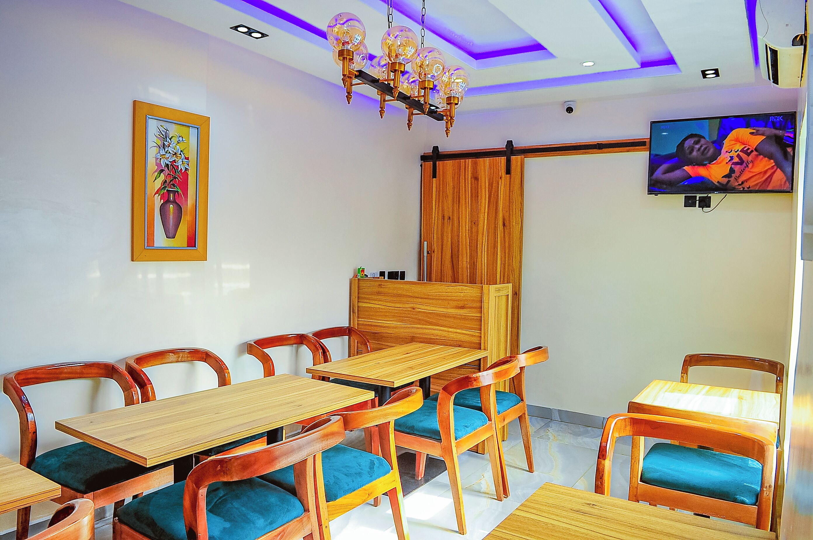 A dining area with wooden tables and chairs, a mounted TV, and a ceiling light fixture.