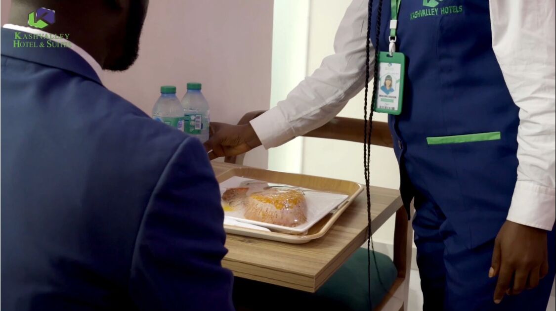 A person handing a tray with a food item to another person in a hotel setting.