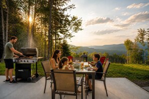 Una familia cena al aire libre cerca de una parrilla, rodeada de árboles y una vista panorámica.