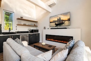 A modern living room with a fireplace, a dining area with a kitchen in the background, and a large window.
