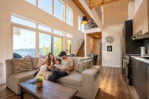 A spacious living room with a large window, a wooden ceiling, and a kitchen area.