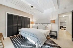 A bedroom with a bed, bedside tables, a wardrobe, a ceiling fan, and a view into another room.