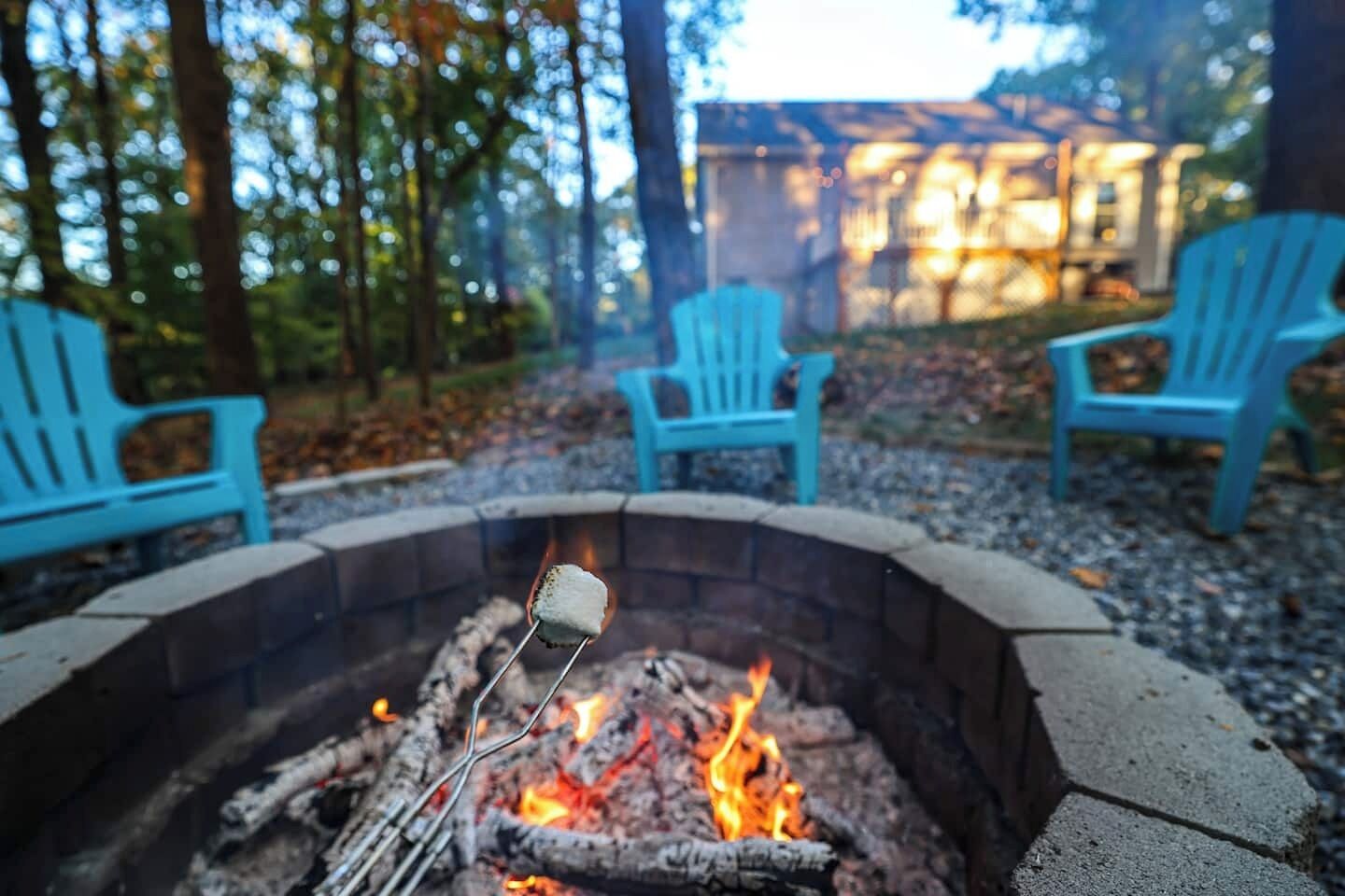 A campfire with a marshmallow on a stick, surrounded by a stone fire pit, with blue chairs and a wooden deck in a wooded area.