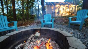 A campfire with a marshmallow on a stick, surrounded by a stone fire pit, with blue chairs and a wooden deck in a wooded area.