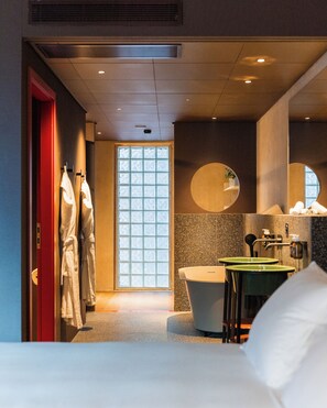 Une chambre d’hôtel avec un lit, une salle de bain dotée d’une baignoire, d’un lavabo et d’un miroir. Des serviettes sont accrochées au mur et une fenêtre laisse entrer la lumière naturelle.