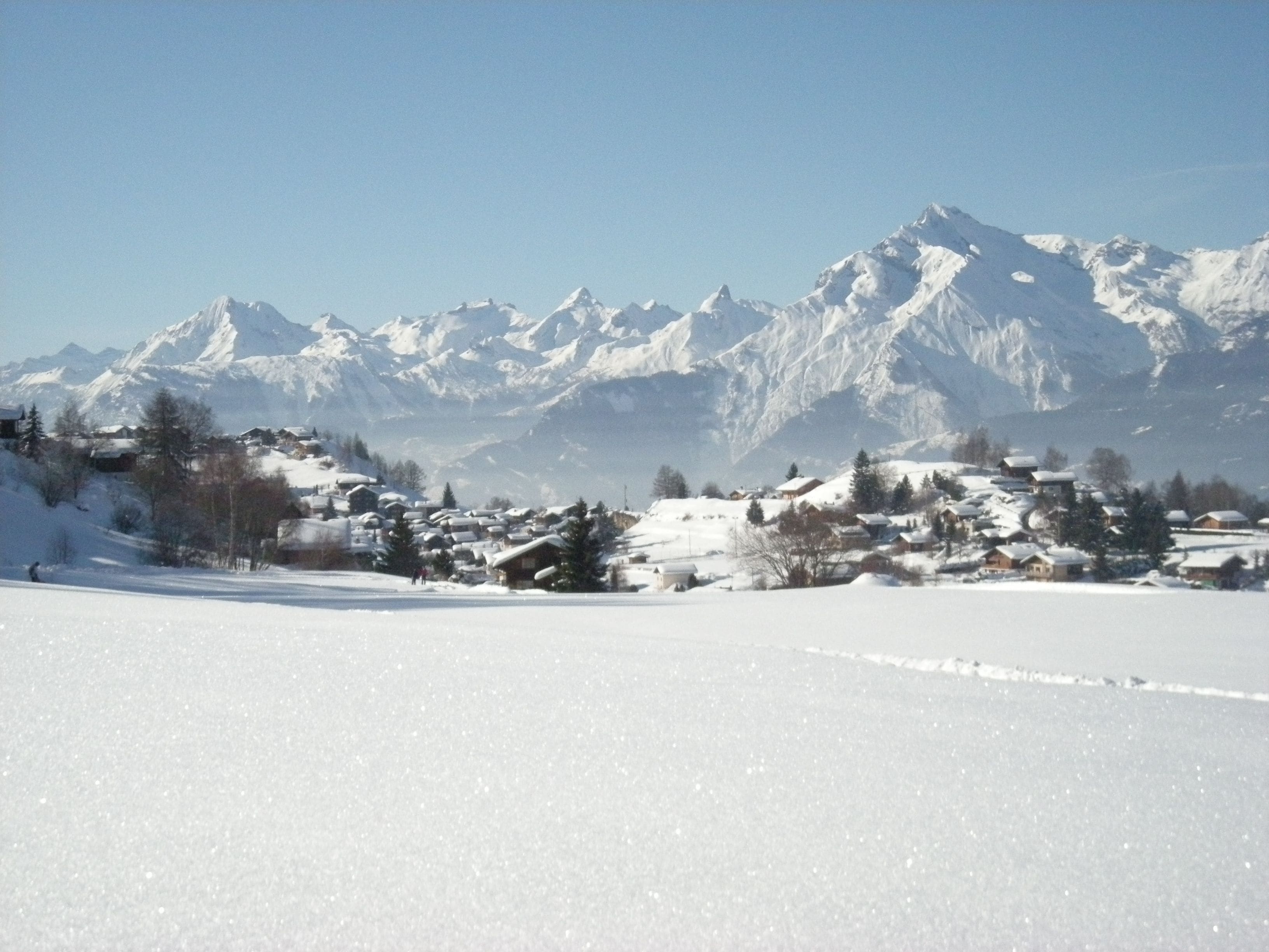 A snowy landscape with a village and snow-covered mountains in the background.