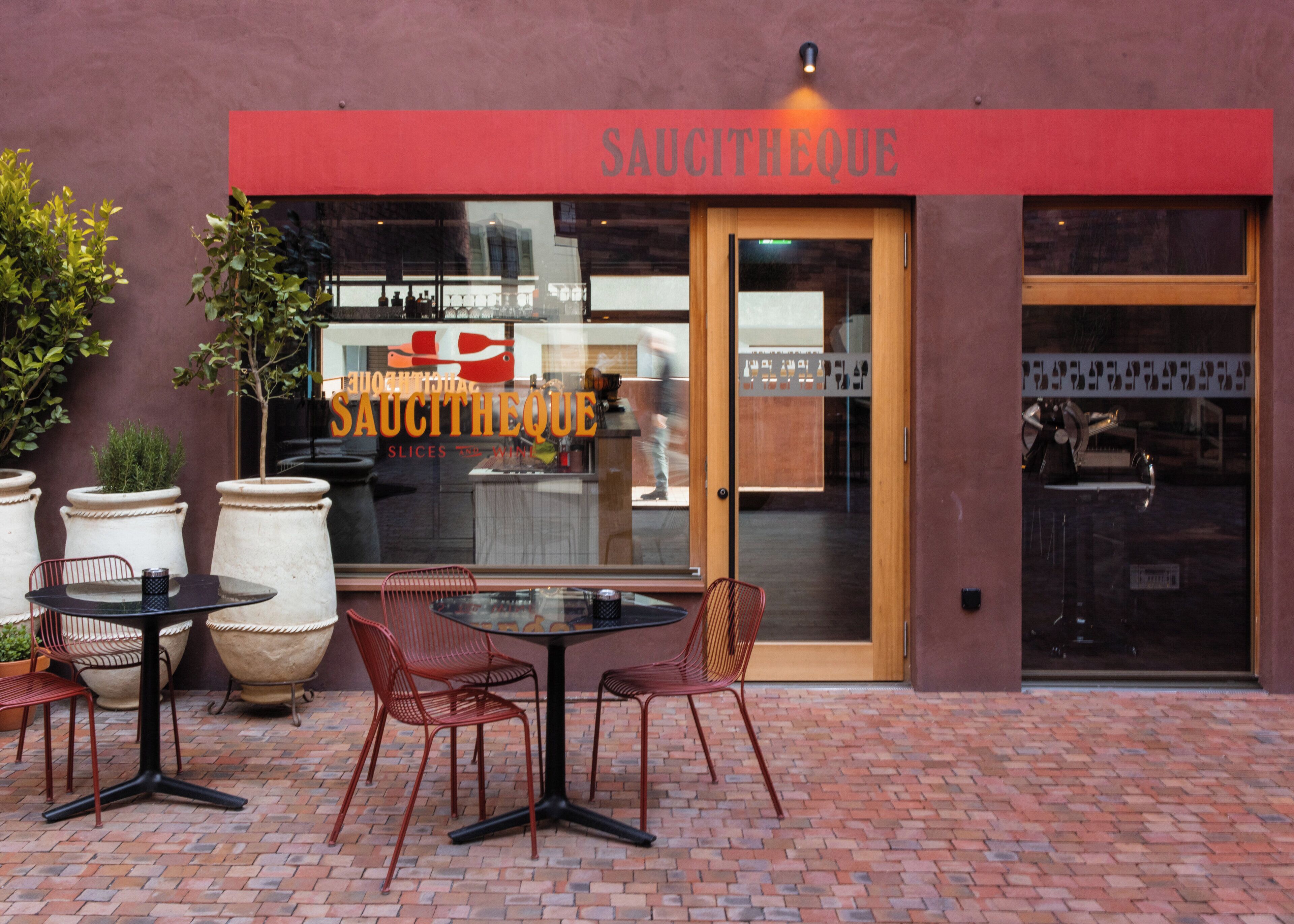 An outdoor seating area with tables and chairs in front of a building with a red awning and a glass door.