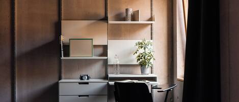 A bedroom with a bed, a desk with a chair, a shelf unit, and a potted plant.
