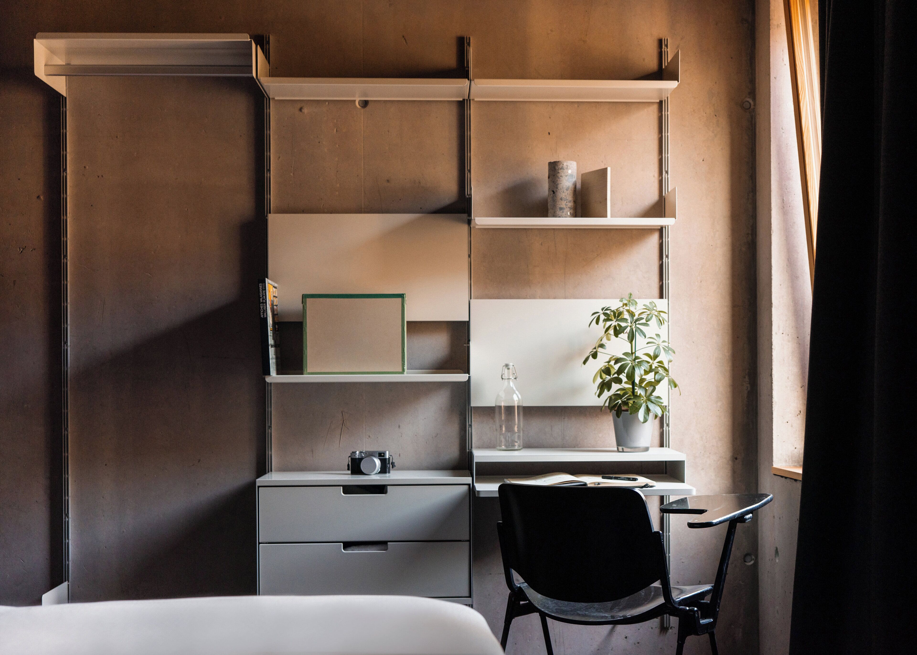 Une chambre à coucher avec un lit, un bureau avec une chaise, une étagère avec des livres et une plante, et une fenêtre avec des rideaux.