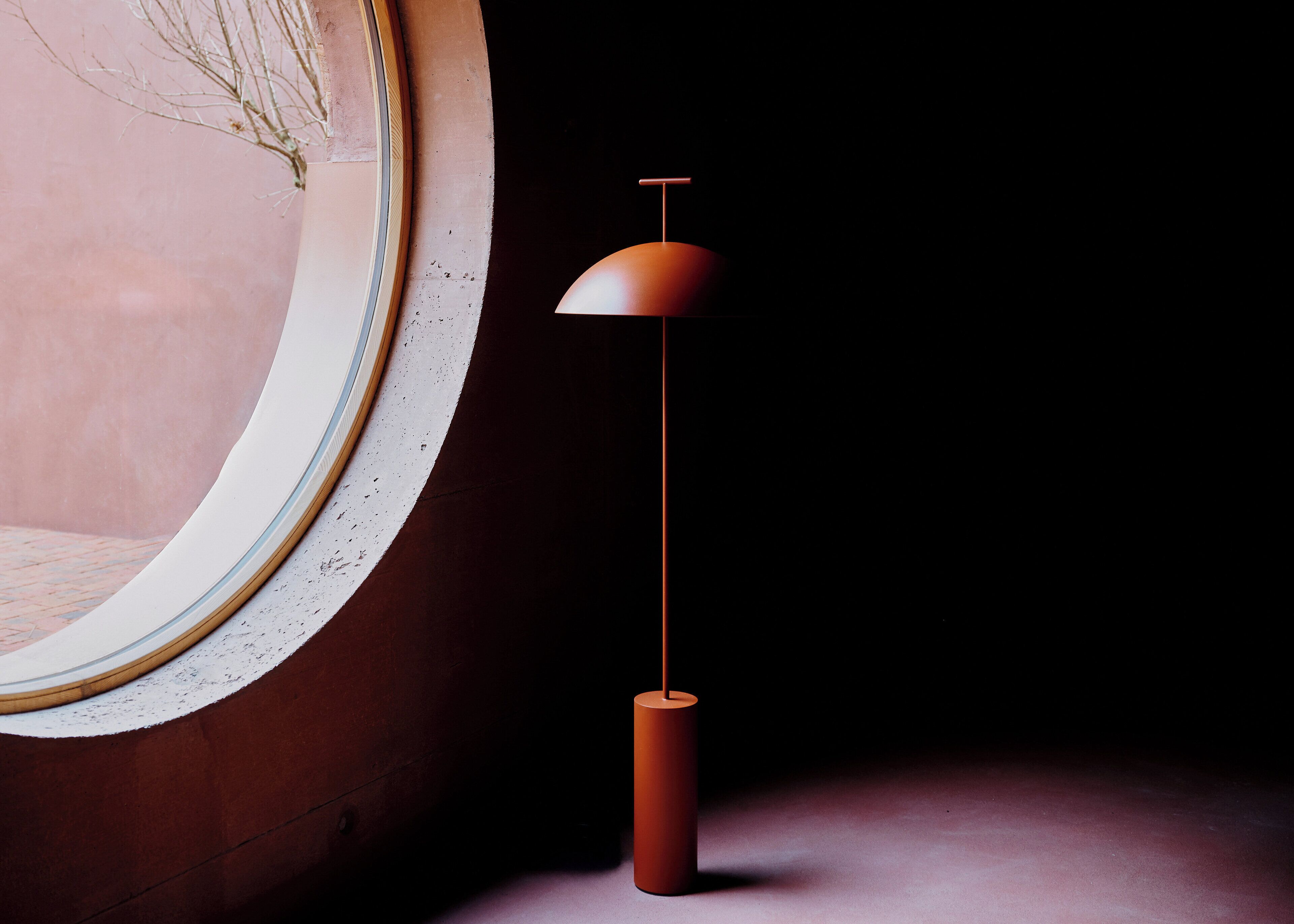 Eine moderne Lampe mit gebogener Basis und einer kuppelförmigen Schirm, platziert auf einem dunklen Boden neben einem großen, runden Spiegel mit strukturierter Rahmung.