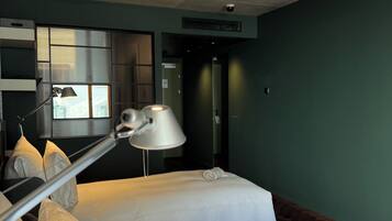 A hotel room with two beds, a desk with a lamp, a window with blinds, and a door.