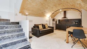 A modern interior with a stone ceiling, a staircase with marble steps, a black leather sofa, a dining table with chairs, and a kitchen area. - An Atypical 1-BR in Place Vendôme Tuileries (Paris)