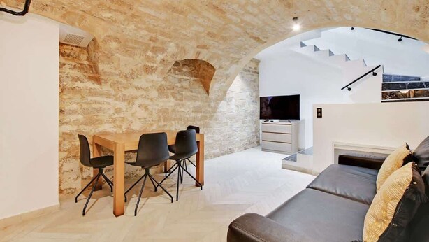 A modern living space with a stone wall, a dining area with a wooden table and black chairs, a grey sofa, and a staircase leading to an upper level. - An Atypical 1-BR in Place Vendôme Tuileries (Paris)