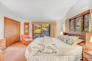A bedroom with a large bed, a wooden wardrobe, a chair, and a window with a view of trees.