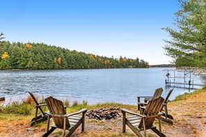 A lakeside view with two wooden chairs facing a fire pit and a dock extending into the water. - Loonsight - PET FRIENDLY (Minocqua)